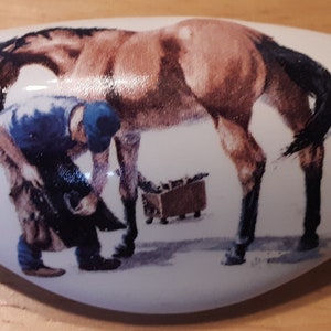 May include: A white ceramic oval-shaped dish with a painted image of a brown horse having its shoe replaced by a blacksmith. The blacksmith is wearing a blue shirt and black pants.