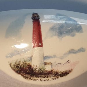 May include: A ceramic, shell-shaped decorative piece featuring a painted image of Barnegat Lighthouse on Long Beach Island, New Jersey. The lighthouse is red and white, set against a sky with clouds. The text "Barnegat Light" is visible.