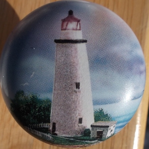 May include: A round, decorative knob featuring a detailed image of a white lighthouse with a red top. The lighthouse is set against a backdrop of a cloudy blue sky and ocean, with green foliage at its base.