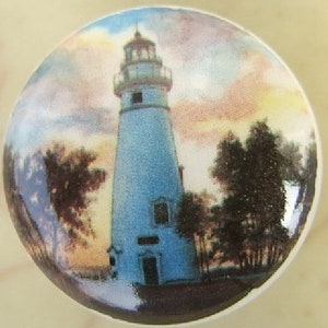 May include: A round, ceramic cabinet knob featuring a detailed illustration of a lighthouse. The lighthouse is light blue with a white top, set against a sunset sky. Trees and water are visible at the base of the lighthouse.