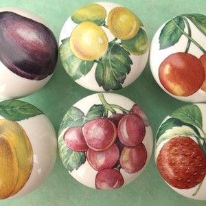May include: Six white ceramic knobs with painted fruit designs. The knobs feature a plum, a yellow apple, a bunch of grapes, two cherries, a lemon, and two strawberries.