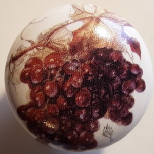 May include: A round, white ceramic cabinet knob featuring a detailed illustration of a cluster of dark red grapes with green leaves. The grapes are rendered in a realistic style, with varying shades of red and purple. The knob is a decorative element for furniture.