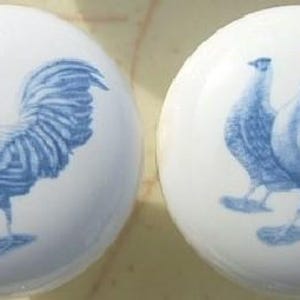 May include: Two white ceramic cabinet knobs with a blue rooster and hen design. The rooster has a detailed tail and a prominent comb. The hen is smaller and stands beside the rooster. The knobs are round and have a glossy finish.