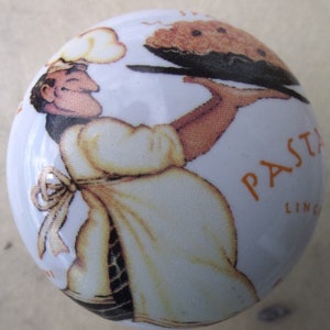 May include: White ceramic cabinet knob featuring a smiling chef in a yellow apron holding a tray of food. The word "PASTA" is printed in orange. A decorative accent for kitchen or dining room furniture.