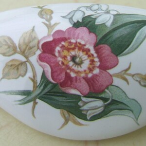 May include: White ceramic drawer pull with a floral design. The design features a large pink flower with a yellow center, green leaves, and smaller white flowers. The pull has a curved shape.