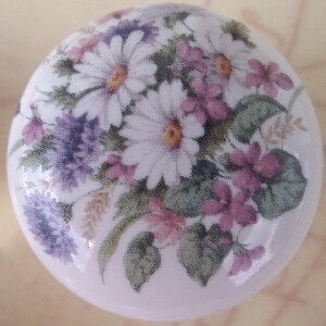 May include: A round, white ceramic knob featuring a floral design. The design includes white daisies with yellow centers, purple and pink flowers, and green leaves. The knob is likely for a cabinet or drawer.