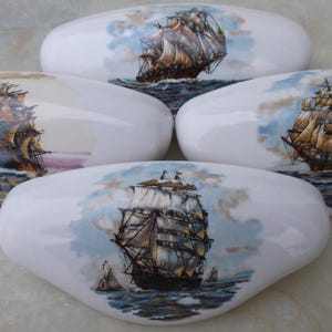 May include: Four white, oval-shaped decorative cabinet knobs. Each knob features a detailed illustration of a tall sailing ship on the ocean, with blue skies and clouds in the background. The ships have brown and tan sails.