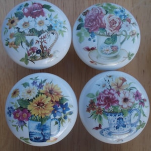May include: Set of four white ceramic cabinet knobs. Each knob features a floral design with a teacup or vase filled with colorful flowers. The flowers include roses, daisies, and other blooms in shades of pink, yellow, and blue.