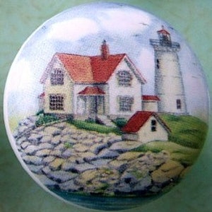 May include: A round decorative knob featuring a coastal scene. The image depicts a white lighthouse and a house with a red roof, set against a blue sky. The knob is likely made of ceramic or porcelain.