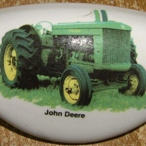 May include: A white ceramic door knob with a green John Deere tractor printed on it. The tractor has a green cab, black wheels, and a yellow front tire.