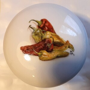 May include: A white, round decorative knob featuring a colorful illustration of chili peppers. The peppers are depicted in shades of red, yellow, and green, with a detailed, realistic style. The knob is likely for a cabinet or drawer.
