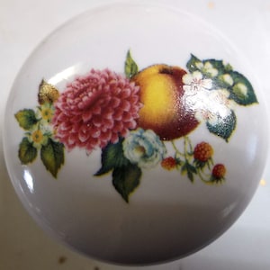 May include: White ceramic cabinet knob with a floral and fruit design. The design includes a large pink dahlia, a yellow and red peach, small blue flowers, and red berries. Green leaves and small white flowers complete the design.