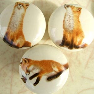 May include: Three white ceramic cabinet knobs with fox illustrations. Each knob features a different pose of a fox in shades of orange, brown, and white. The knobs are round and have a glossy finish, suitable for furniture or decorative projects.