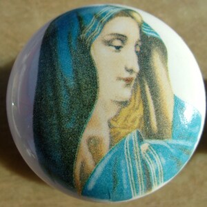 May include: A round, white ceramic knob featuring a portrait of a woman with a blue head covering. The portrait has a vintage aesthetic, with soft colors and delicate details. The knob is likely for a cabinet or drawer.