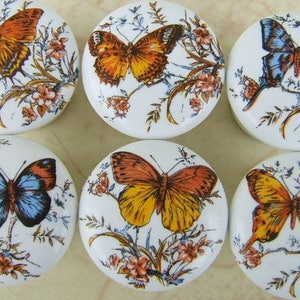 May include: Six white ceramic cabinet knobs with butterfly designs. Each knob features a different butterfly in shades of orange, yellow, and blue, with floral accents. The knobs are round and have a glossy finish.