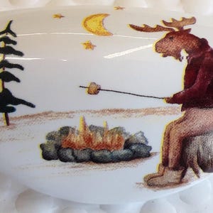 May include: White ceramic golf ball marker with a whimsical illustration of a moose roasting a marshmallow over a campfire. The scene includes pine trees, a crescent moon, and stars. The moose is wearing a maroon jacket.
