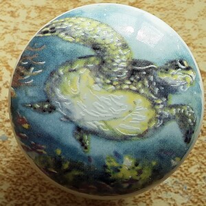 May include: A round decorative knob featuring a sea turtle swimming in a turquoise ocean. The knob has a glossy finish and a detailed illustration of the turtle with yellow, green, and white accents. Coral and other sea life are visible in the background.