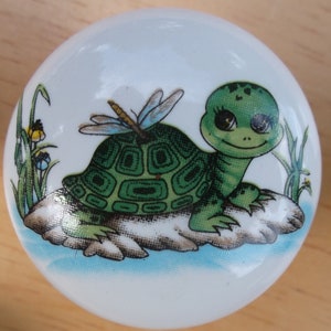 May include: A white ceramic knob with a green turtle and dragonfly design. The turtle has big eyes and is sitting on a rock. The dragonfly has blue wings and is flying above the turtle.