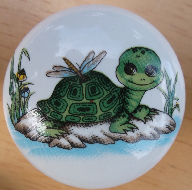 May include: A white ceramic knob with a green turtle and dragonfly design. The turtle has big eyes and is sitting on a rock. The dragonfly has blue wings and is flying above the turtle.