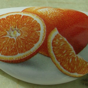 May include: A white ceramic dish with a painted image of three orange slices. The oranges are a vibrant orange color with a yellow center. The dish is oval shaped and has a smooth, glossy finish.