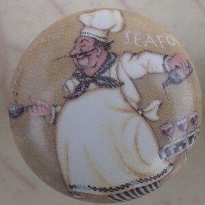 May include: A round fabric button featuring a cartoon chef pouring wine. The chef wears a white hat and coat, with the word "SEAFOOD" visible. The background is a light tan color.