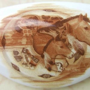 May include: White ceramic drawer pull featuring a sepia-toned illustration of three horses. The horses are depicted with detailed manes and faces, surrounded by Native American-inspired symbols. The pull has a curved, elongated shape.