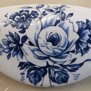 May include: A white ceramic oval dish with a blue floral design. The design features a large rose with smaller flowers and leaves.