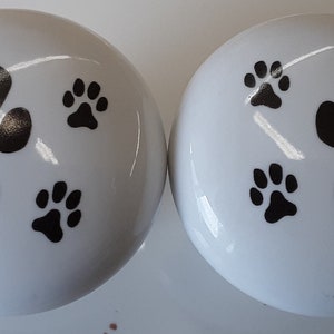 May include: Two white ceramic knobs with black paw print designs. Each knob has four paw prints, one large and three small.