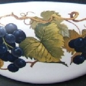 May include: White, oval-shaped decorative dish with a painted design of dark blue grapes, green and yellow leaves, and brown vines. The grapes are clustered together, and the leaves have detailed veining. The dish has a smooth, glossy finish.