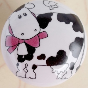 May include: A white ceramic cabinet knob featuring a cartoon cow design. The cow has black spots, a pink bow, and a small bird. The knob is round and likely used for furniture or decorative purposes.