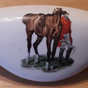 May include: White ceramic door knob with a brown horse and a person in a red jacket and white pants. The horse is wearing a brown saddle.
