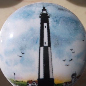 May include: A round, white ceramic knob featuring a lighthouse design. The lighthouse is black and white, set against a blue sky with clouds and flying birds. The base of the knob shows a small building and a grassy area.
