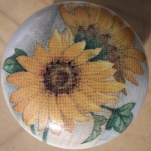May include: A round, white ceramic cabinet knob featuring a detailed image of two sunflowers. The sunflowers have yellow petals and dark brown centers, with green leaves and stems. The knob has a glossy finish.