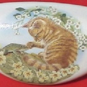 May include: White ceramic decorative piece with a painted image of a tabby cat interacting with a frog amidst flowers and foliage. The cat is brown with dark stripes. The flowers are yellow and white.