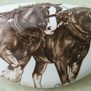 May include: White oval decorative knob featuring a sepia-toned illustration of two draft horses in harness. The horses are depicted in motion, with detailed harnesses and bridles. A charming accent for cabinets or drawers.