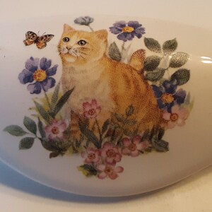 May include: A white ceramic knob with a painted design of a ginger cat sitting in a garden with flowers and a butterfly. The cat is surrounded by pink, purple, and blue flowers.