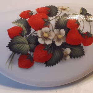 May include: White ceramic drawer pull with a vibrant strawberry design. The strawberries are a bright red with green leaves and small white flowers. The pull is oval-shaped and has a glossy finish, suitable for kitchen or furniture decor.