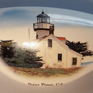 May include: A white, oval-shaped decorative item featuring a painted image of the Point Pinos lighthouse in California. The lighthouse is white with a black top and red roof, surrounded by green trees. The text "Point Pinos, CA" is at the bottom.