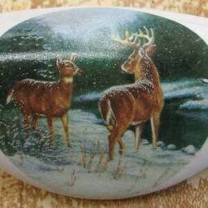 May include: White ceramic drawer pull with a winter scene featuring two deer in a snowy forest. The deer are brown and white with antlers. The background is a green forest with snow on the ground.