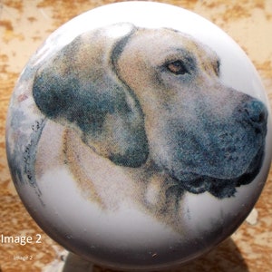 May include: A white, round cabinet knob featuring a detailed portrait of a dog's head in profile. The dog has tan and gray fur, with a focused gaze. The knob is likely ceramic or porcelain.