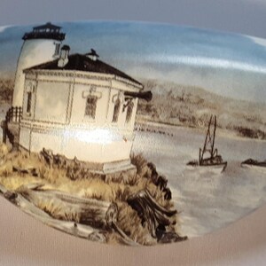 May include: A decorative, oblong-shaped object with a painted coastal scene. The artwork depicts a white lighthouse on a rocky shore, sailboats on the water, and a blue sky. The object has a glossy finish.