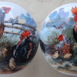 May include: Two ceramic knobs with a rooster and hen design. The knobs feature a rooster with a red comb and a hen with a brown and white patterned body. Both knobs have a green and brown background with a fence and other chickens.