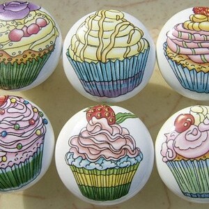 May include: Six ceramic cupcake drawer knobs with colorful frosting and sprinkles. The cupcakes are decorated with different designs, including a swirl, a strawberry, and a cherry.