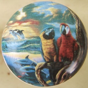 May include: A ceramic knob with a colorful image of a tropical scene. The scene includes two parrots perched on a branch, dolphins swimming in the ocean, and a sunset in the background.