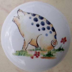 May include: A white ceramic knob with a cartoon image of a spotted pig. The pig is white with black spots and is standing on a green patch of grass with red flowers.