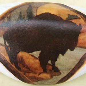 May include: An oval-shaped decorative stone featuring a dark brown bison silhouette within a landscape scene. The bison stands on a tan surface, framed by a brown border against a white background. The artwork evokes a rustic, nature-inspired theme.