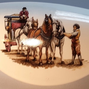 May include: A white ceramic oval-shaped dish with a painted scene of a horse-drawn carriage with three horses and two people. The horses are brown and white, and the carriage is red and black.