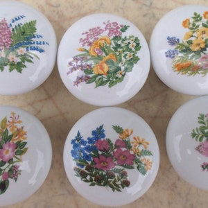 May include: Six white ceramic cabinet knobs with floral designs. Each knob features a different arrangement of colorful flowers, including pink, yellow, blue, and purple blooms, set against a white background. The knobs are round and have a glossy finish.