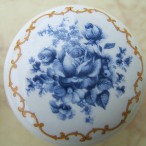 May include: A white ceramic cabinet knob with a blue floral design. The knob features a detailed illustration of blue roses and foliage. A gold-colored decorative border frames the floral pattern.