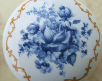 May include: A white ceramic cabinet knob with a blue floral design. The knob features a detailed illustration of blue roses and foliage. A gold-colored decorative border frames the floral pattern.